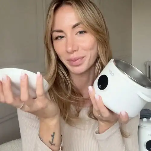 A woman with blonde hair holds a white electric bowl with a stainless steel interior and its lid.