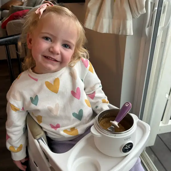 A smiling toddler in a heart-patterned sweatshirt sits in a high chair with a bowl of food.