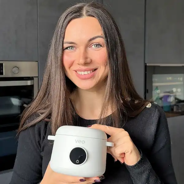 A smiling woman with long brown hair holds a small, white Veo portable cooker in a kitchen.