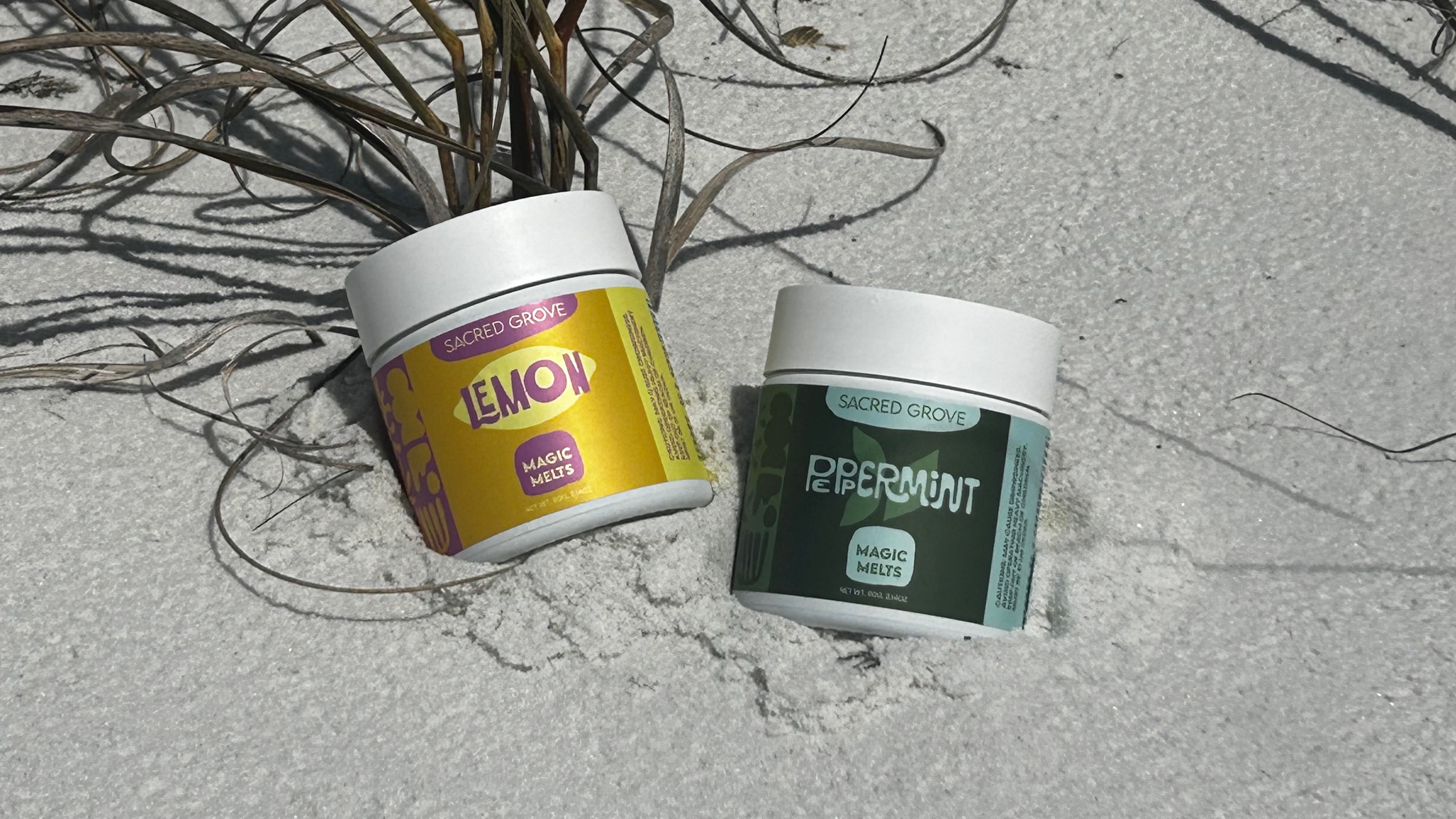 Two jars of Sacred Grove Magic Melts, lemon and peppermint flavors, resting on white sand with dried grass.