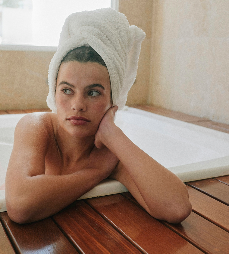 A woman with a towel on her head leans on the side of a bathtub, looking away.