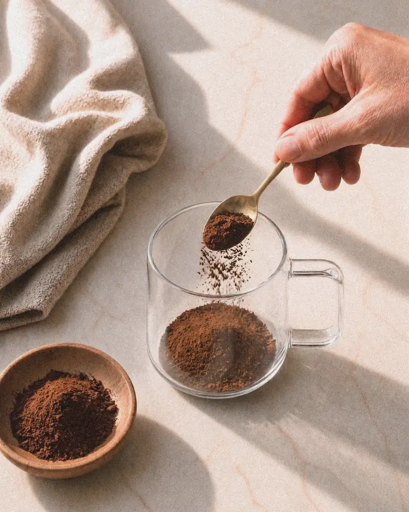 A person pours milk into a glass mug of a chocolate drink while stirring it.