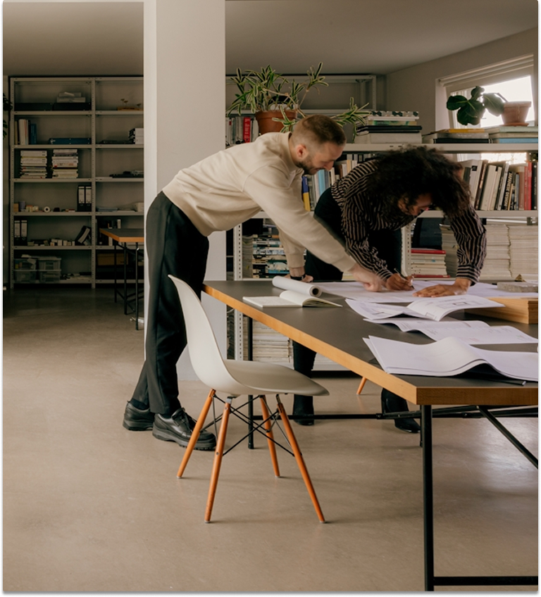 Two people lean over a large table in a modern office, reviewing architectural plans.