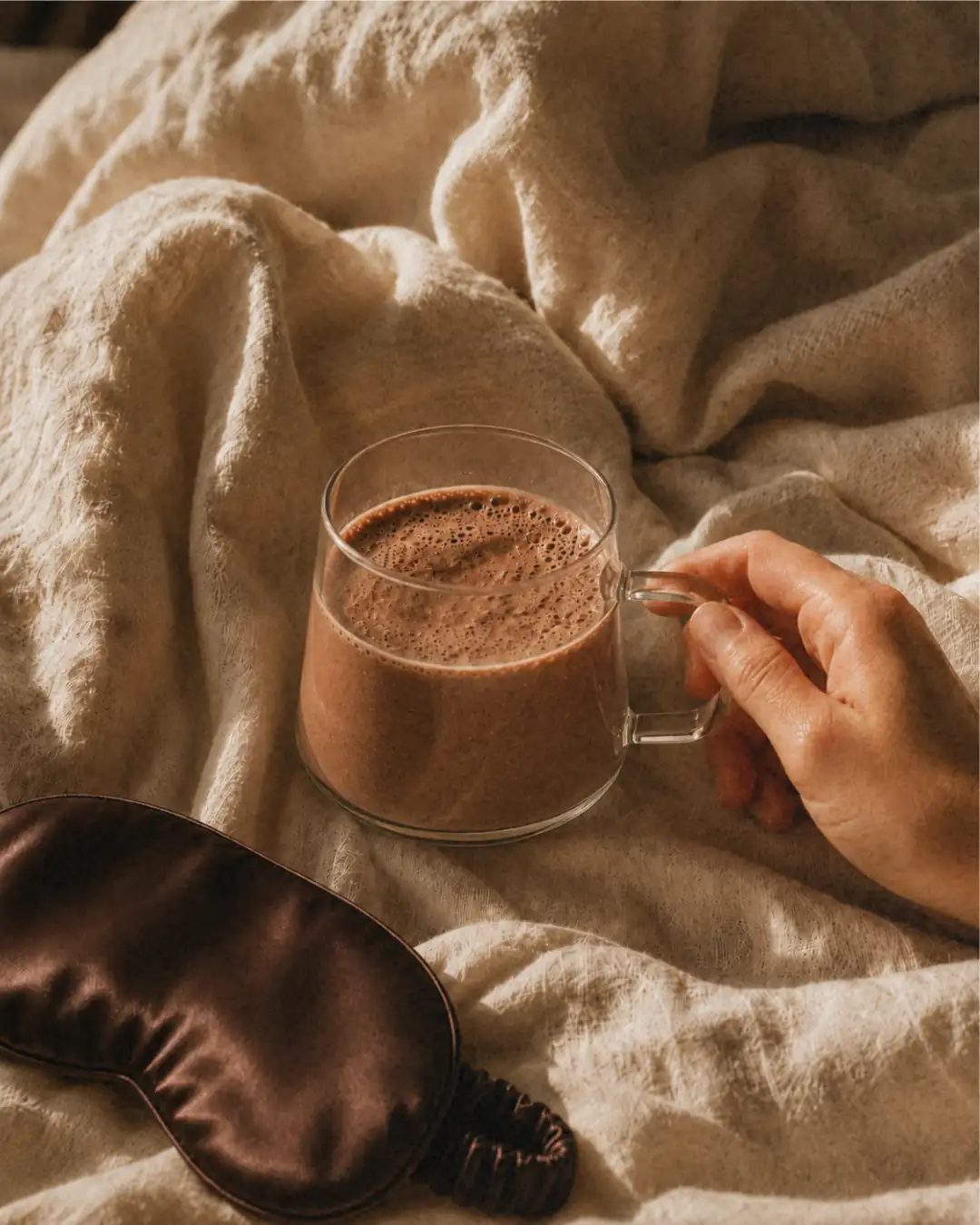 A hand holding a clear mug of a frothy brown drink on a rumpled blanket next to a sleep mask.