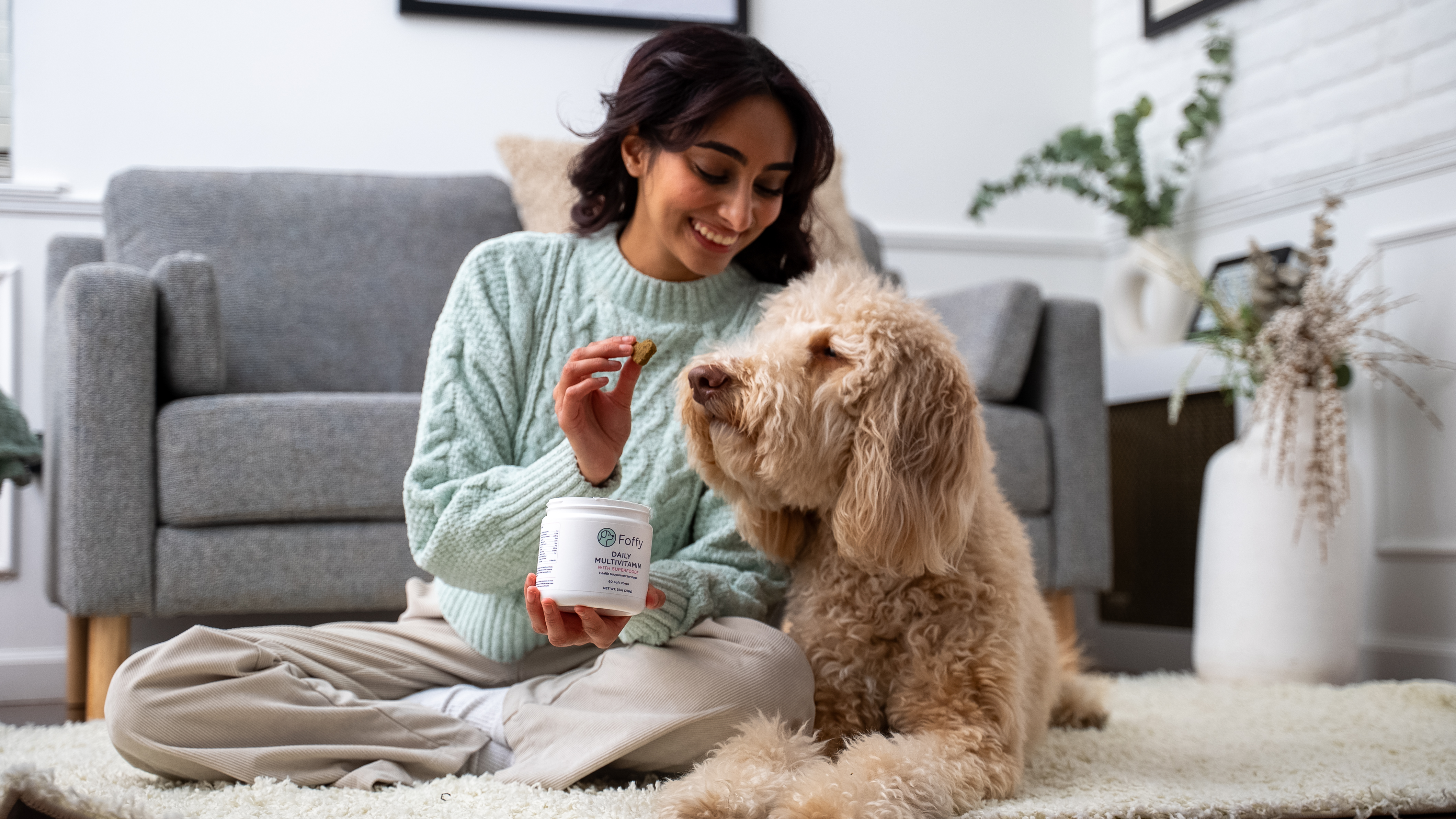 A smiling man in glasses holds a chewable supplement for a small brown dog.