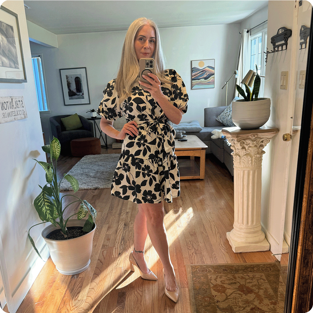 A woman with long grey hair takes a mirror selfie in a floral dress and heels.