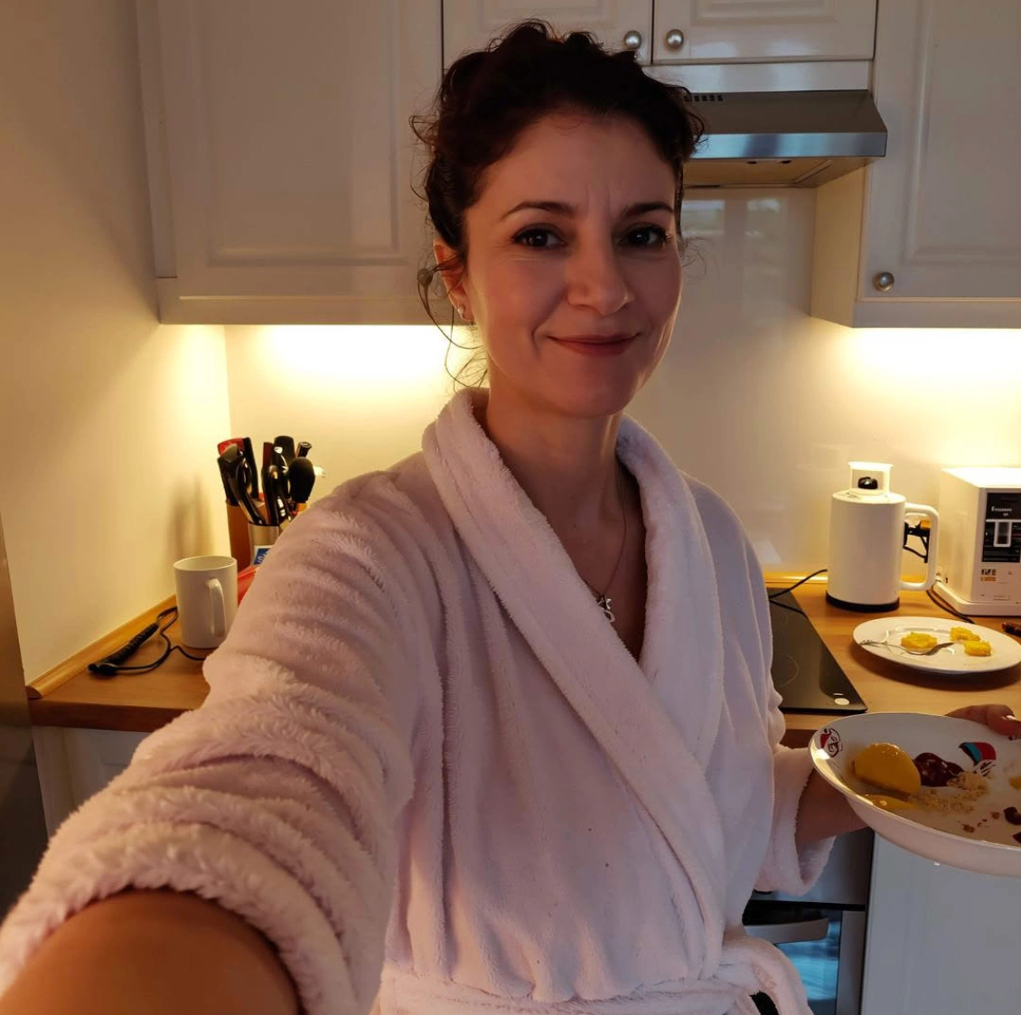 A smiling woman in a pink bathrobe holds a plate of food in a kitchen.