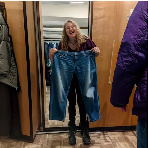 A laughing woman holds up a pair of oversized blue jeans in what appears to be a store.