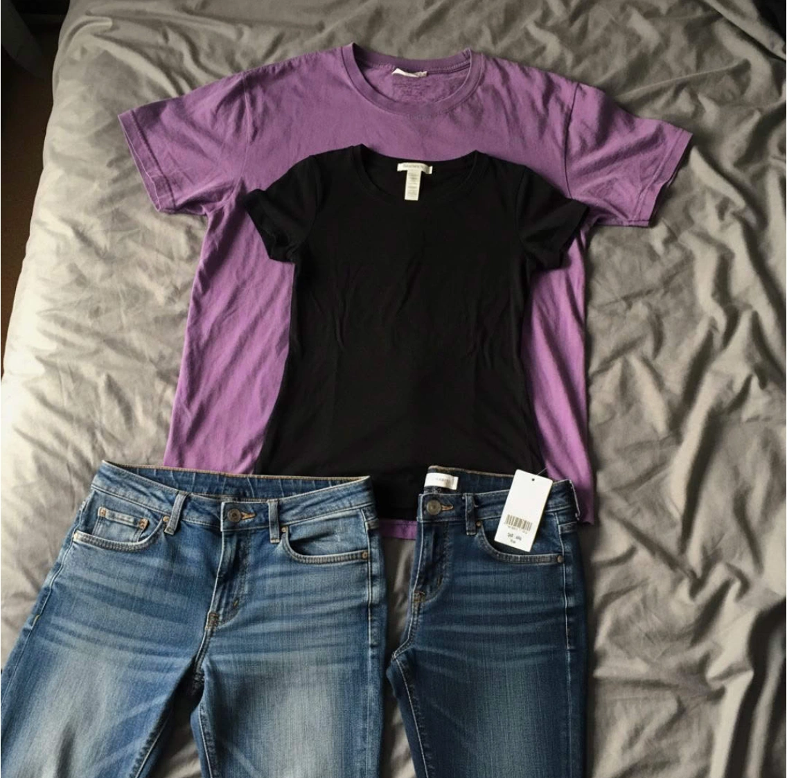 Two t-shirts and two pairs of jeans are laid out on a gray, wrinkled bedspread.