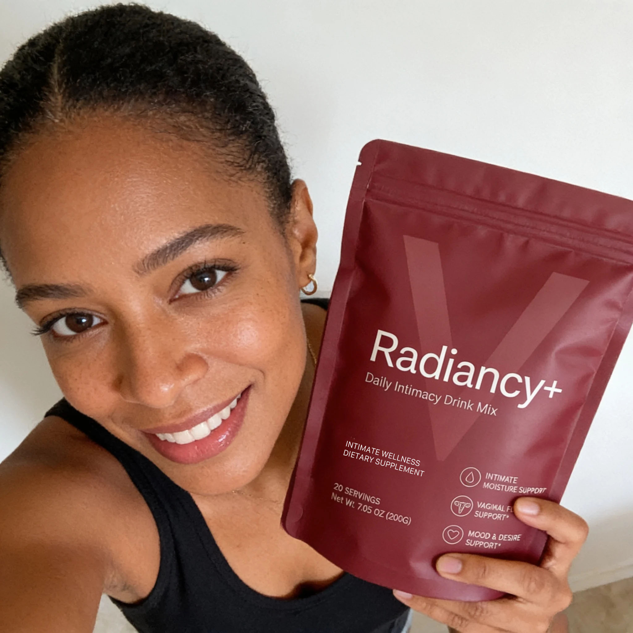 A smiling woman holds up a maroon pouch of Radiancy+ Daily Intimacy Drink Mix dietary supplement.