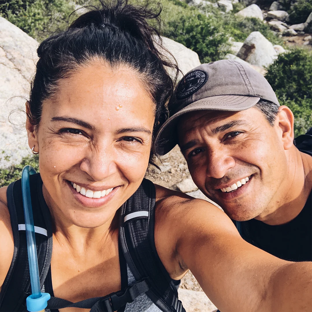A close-up selfie of a smiling man and woman outdoors in a rocky, brushy area.