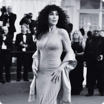 A black and white photo of Cher in a glamorous, beaded gown and shawl, posing on a red carpet.