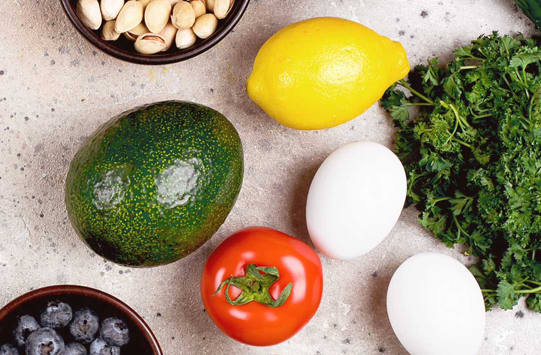 Various foods: pistachios, lemon, avocado, eggs, tomato, parsley, and blueberries on a surface.