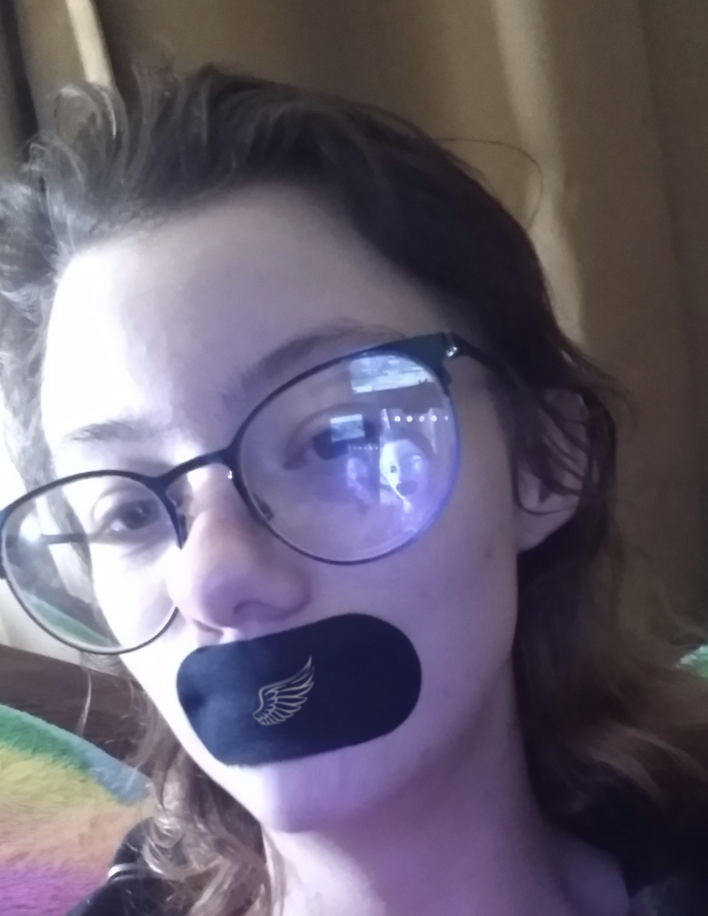 Person with glasses and a black patch over mouth, indoors.
