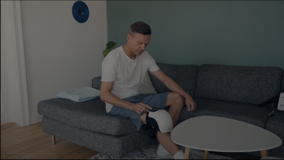 Man sitting on a couch using a knee therapy device.