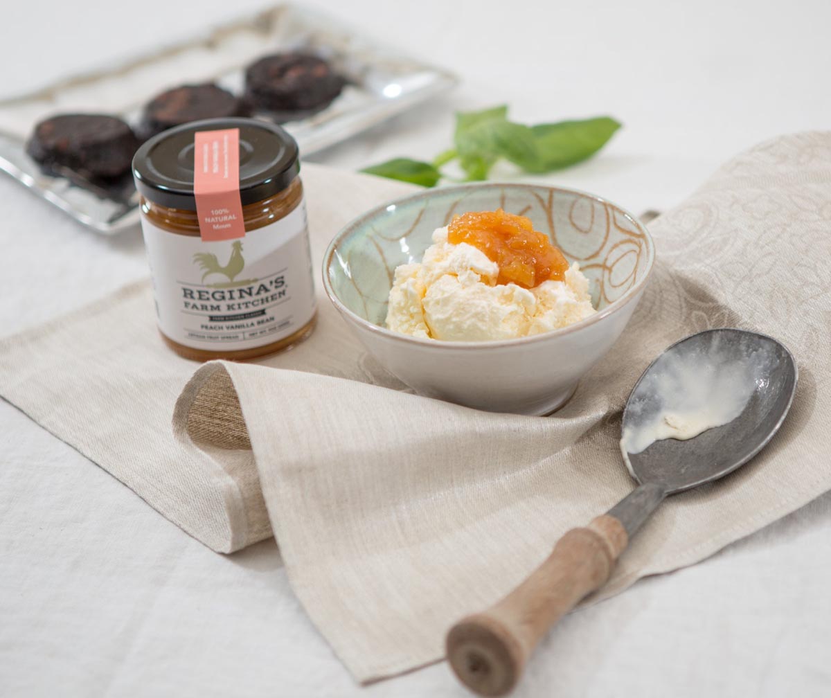 Ice cream with peach topping in a bowl, beside a jar and a wooden-handled scoop.
