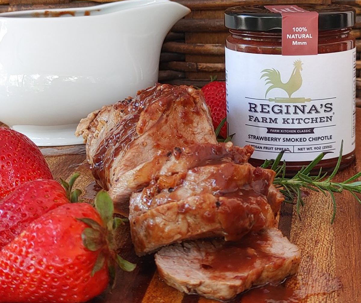 Sliced pork tenderloin with strawberry smoked chipotle sauce, surrounded by fresh strawberries and a sauce jar.
