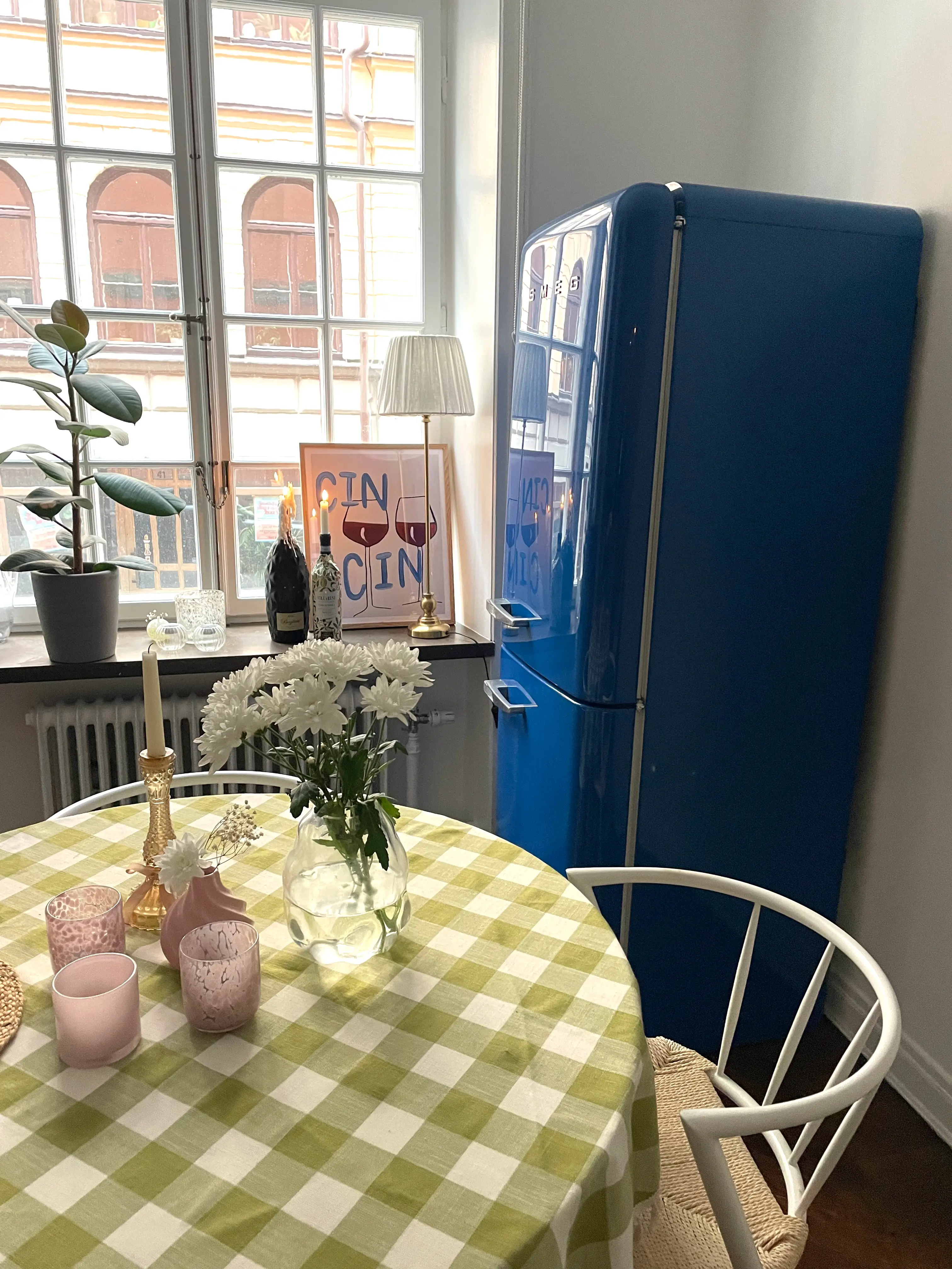 Kitchen with blue fridge, checkered tablecloth, flowers, and window view.
