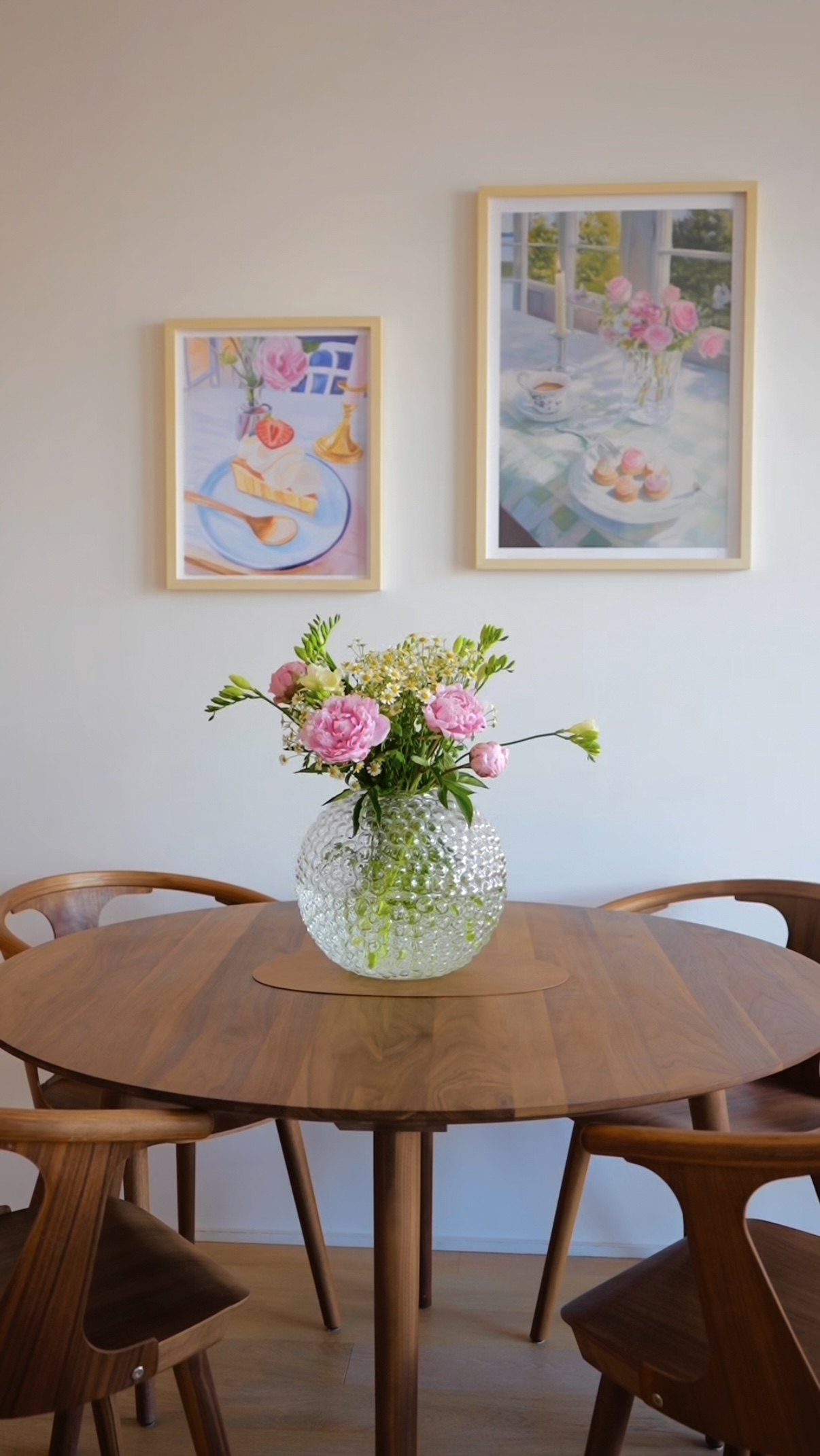 Wooden table with a glass vase of pink flowers, two framed paintings on the wall.