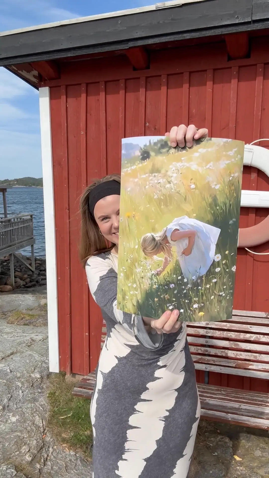 Person holding a painting of a girl in a field, standing by a red building.