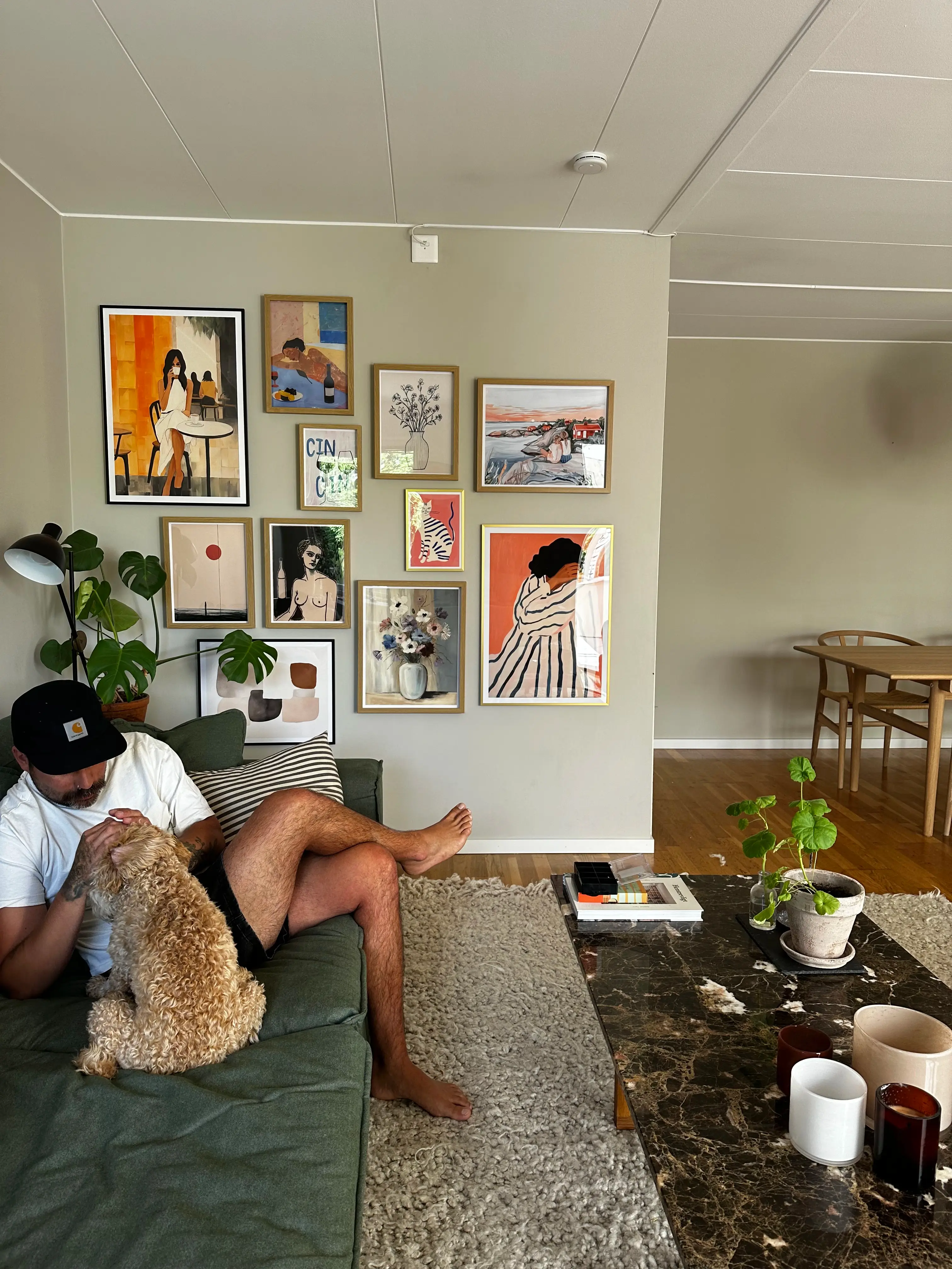 Man sitting on a couch with a dog, surrounded by framed art on the wall.