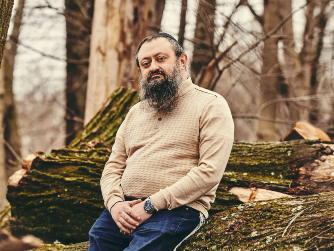 Man with a beard sitting on a fallen tree in a forest.