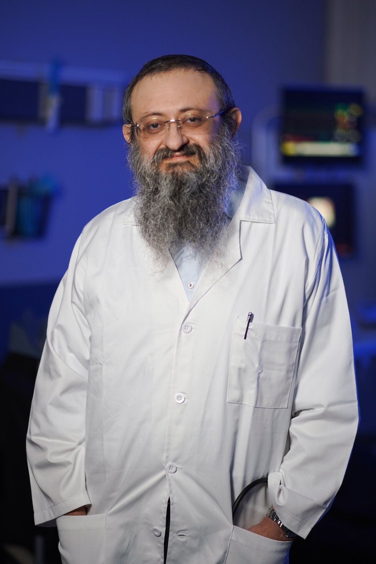 Man with a beard in a white lab coat, standing indoors.