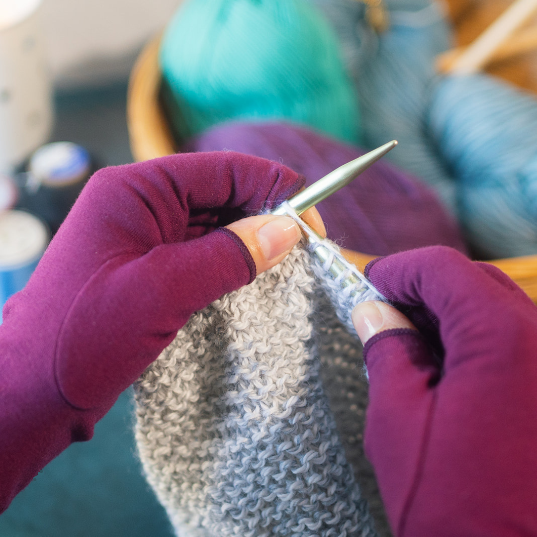 Hands in purple gloves knitting with gray yarn and needles.