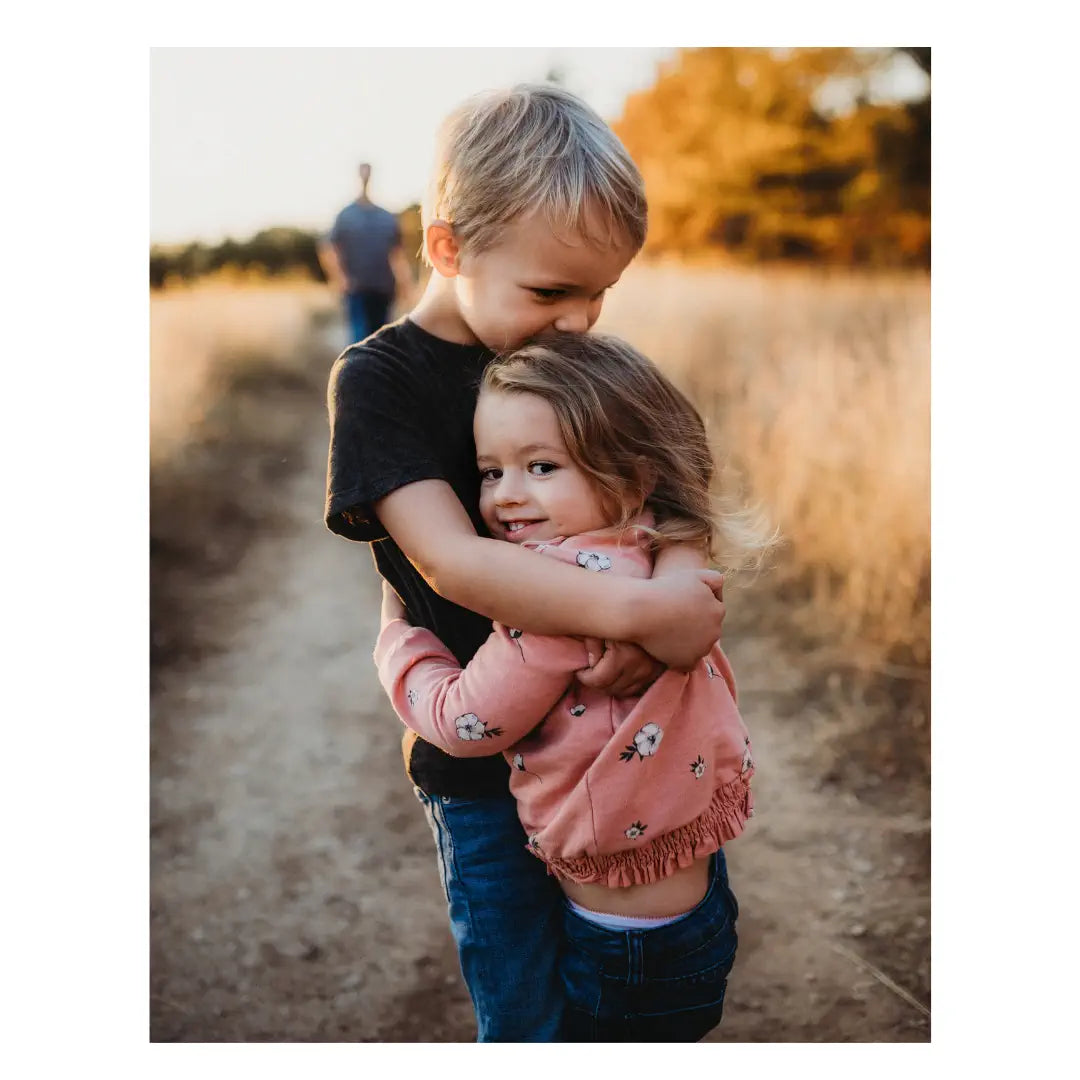 Two children hugging on a dirt path with autumn trees in the background.
