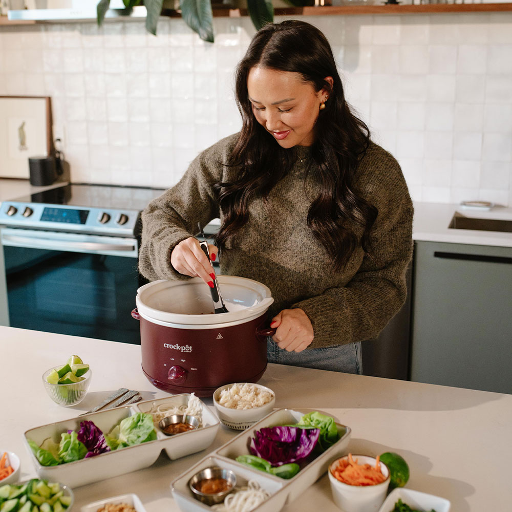 A woman stirs a red Crock-Pot on a kitchen counter surrounded by bowls of fresh food ingredients.