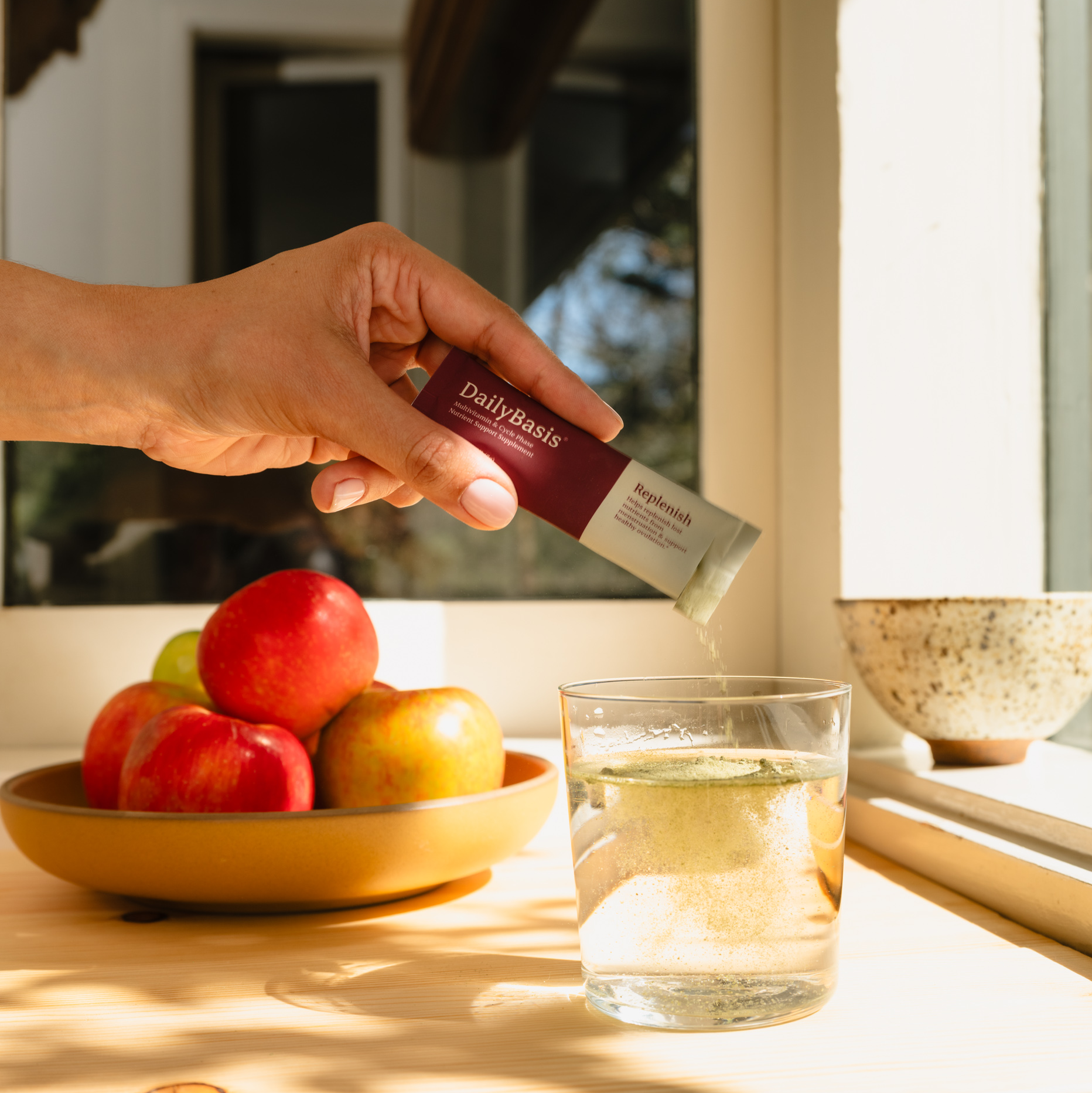 A person pours a DailyBasis supplement packet into a glass of water on a sunlit wooden counter.