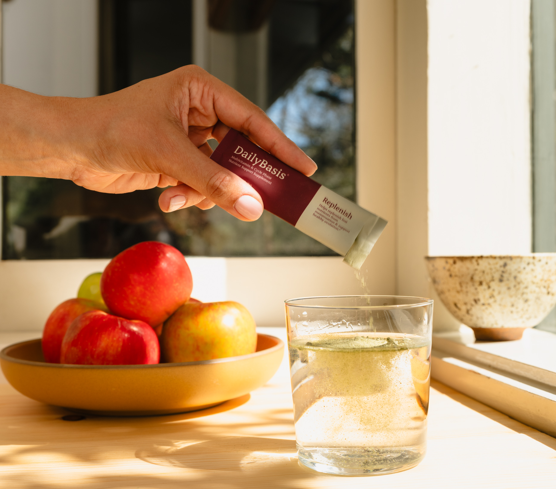 Hand pouring DailyBasis Cycle Routine Replenish supplement stick pack into a glass of water with fresh apples in the background