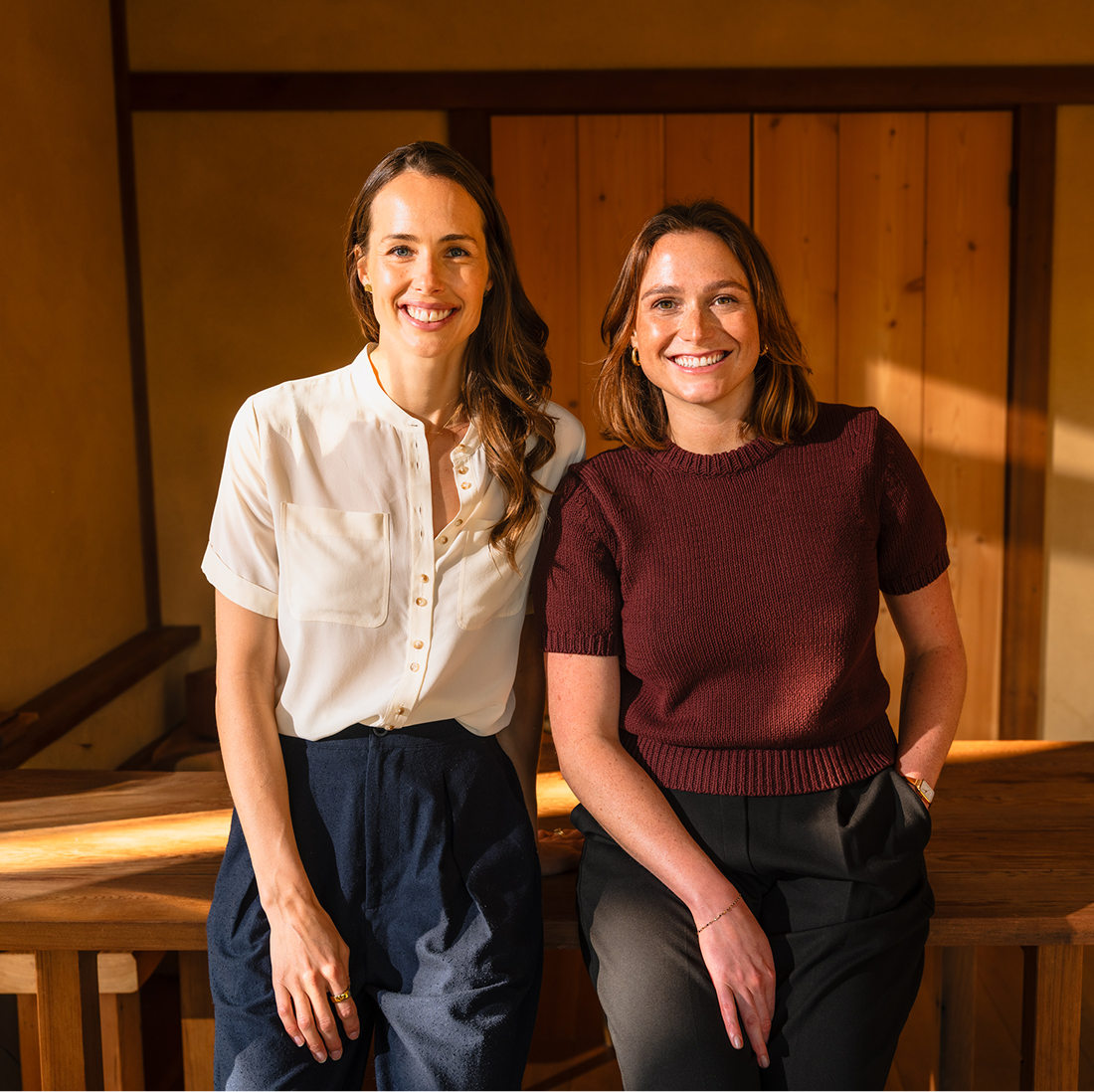 Two women smile at the camera while leaning against a wooden table in a warmly lit room.