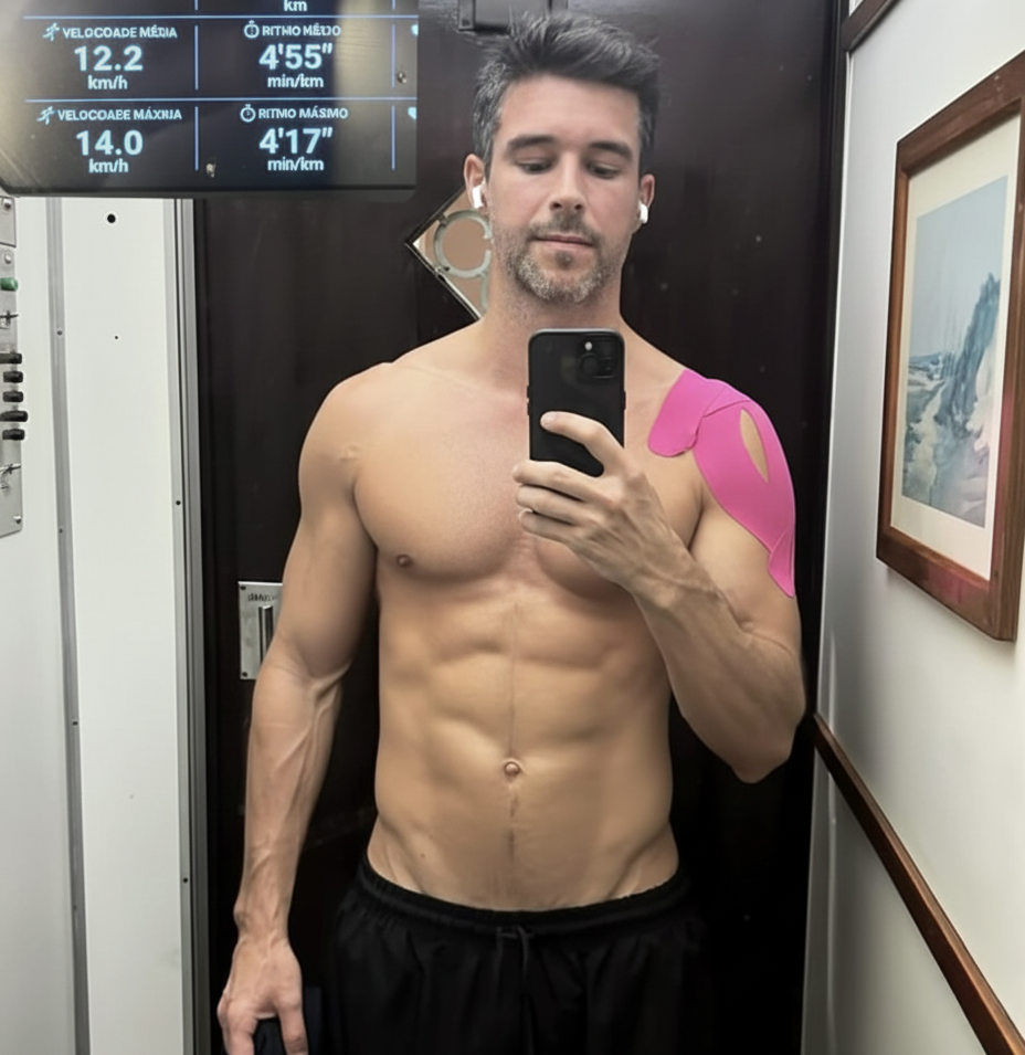 Man taking a shirtless mirror selfie, wearing earbuds and pink shoulder tape.