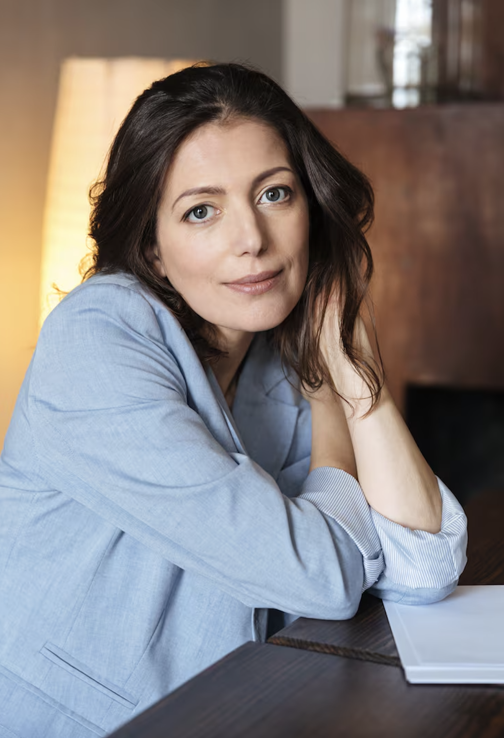 A woman with dark hair in a light blue blazer sitting at a wooden table, looking at the camera.