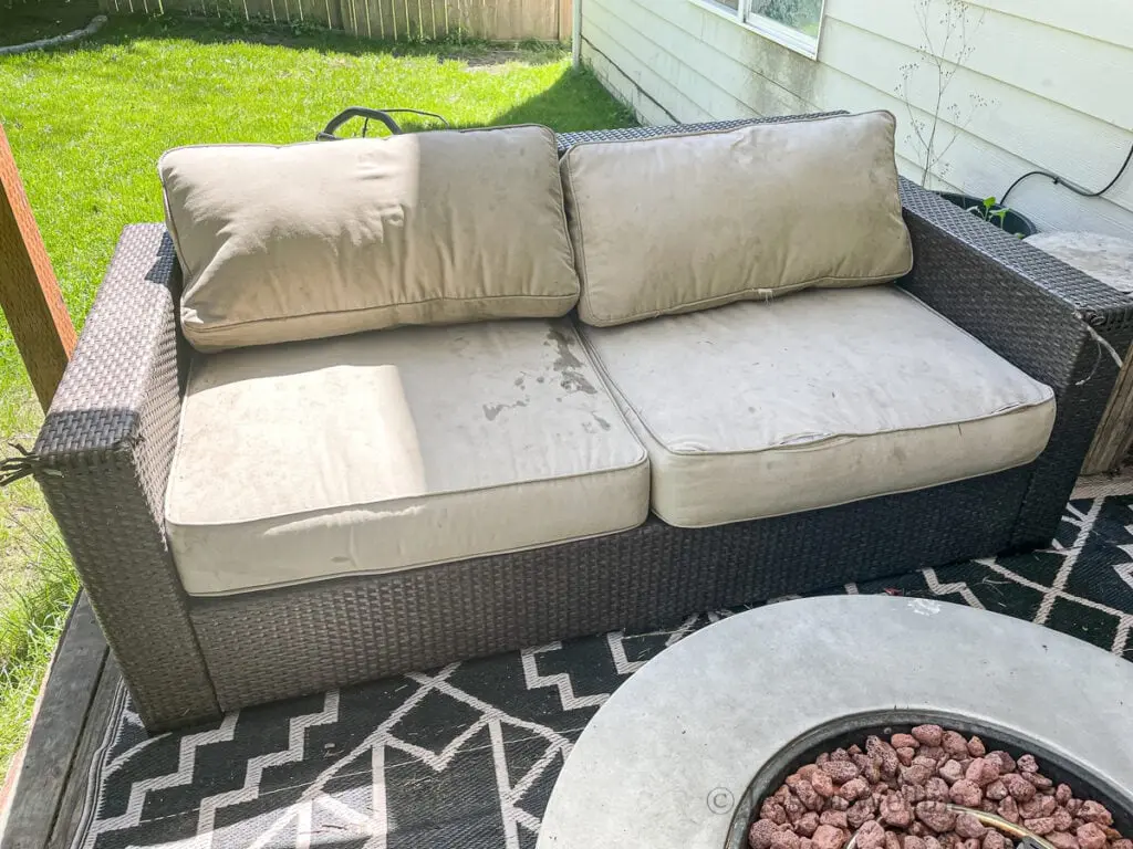 A dirty wicker outdoor loveseat with stained beige cushions sits on a patio next to a lawn.