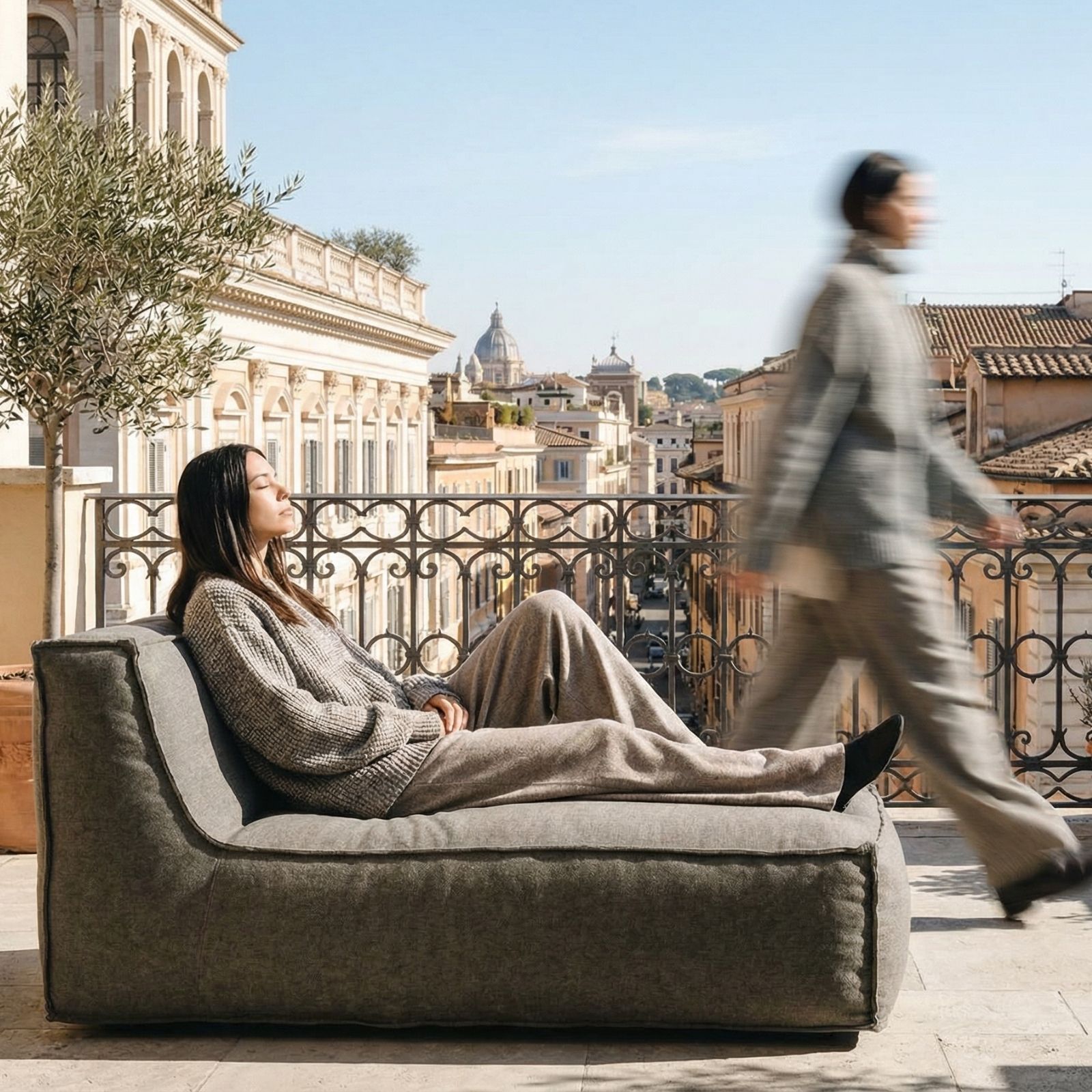 A woman relaxes on a grey lounge chair on a sunny city balcony as another person walks by.