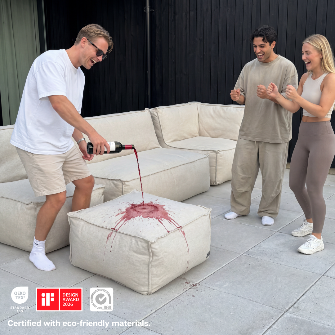 A man pours red wine onto a light-colored ottoman while two people watch and laugh.