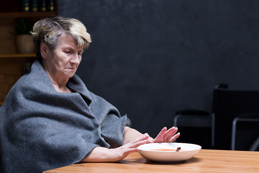 Elderly person in a gray shawl sitting at a table with a bowl.