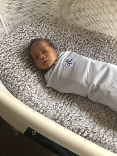 A newborn baby sleeps swaddled in a light blue blanket inside a bassinet with a grey fleece lining.