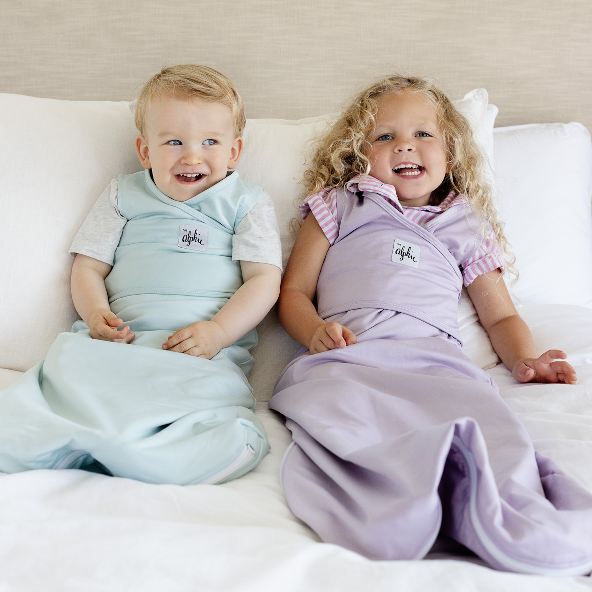 Two children in pastel sleep sacks sitting on a bed, smiling.