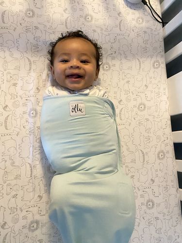A smiling baby swaddled in a light blue blanket lies in a crib on a patterned sheet.