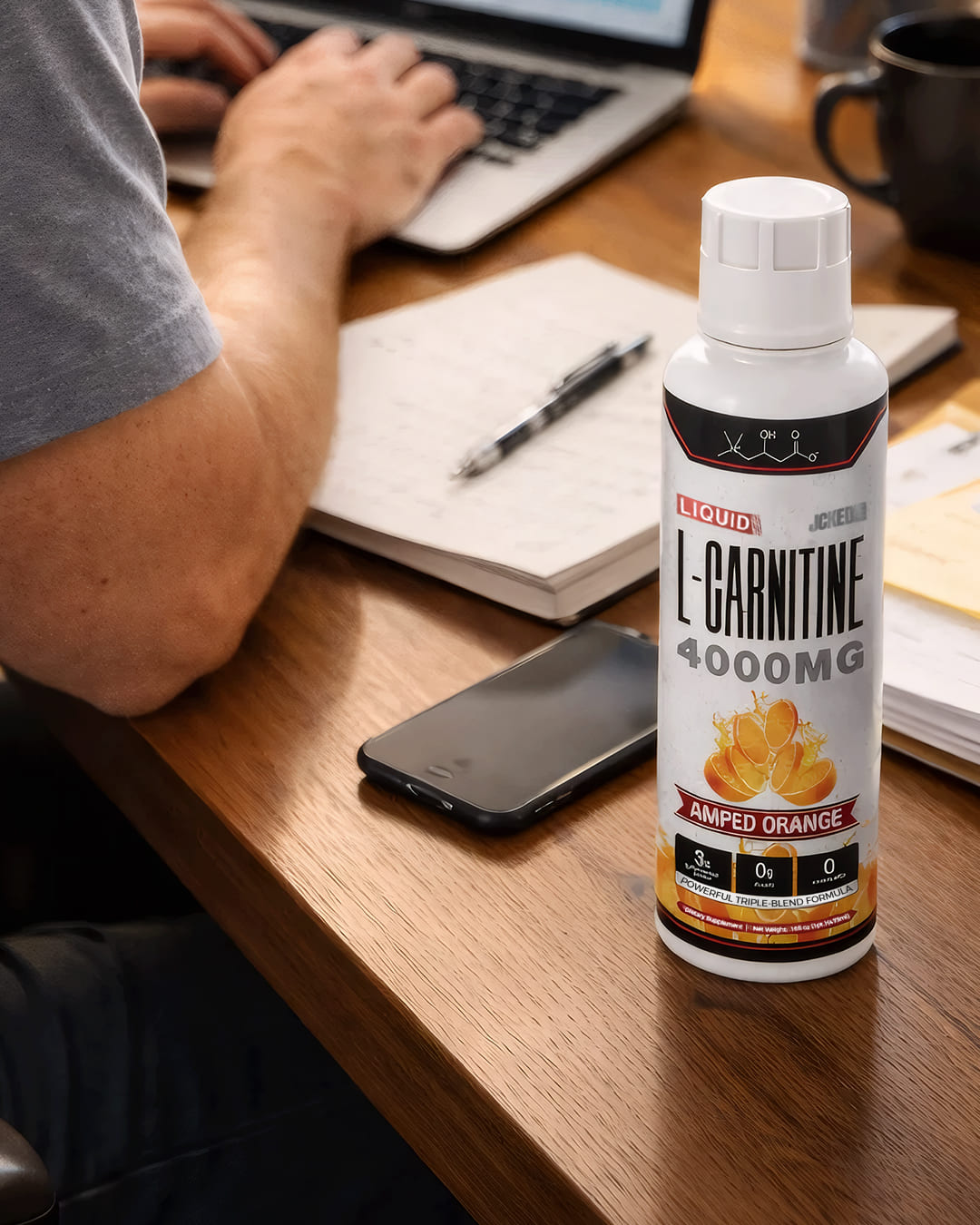A bottle of L-Carnitine supplement on a desk next to a person working on a laptop.