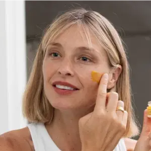 A blonde woman with a white tank top applies an orange-colored product to her cheek with her finger.
