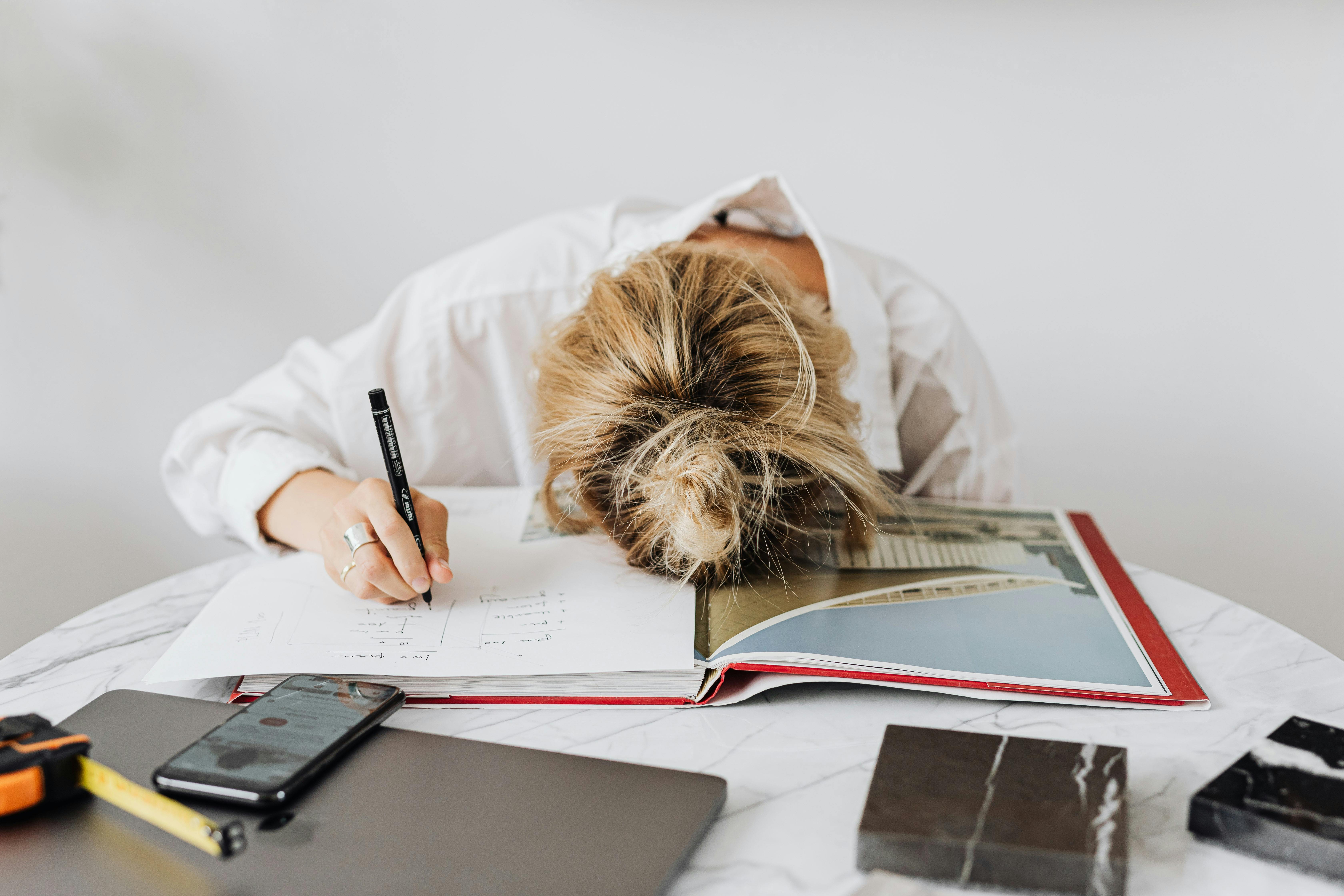 Person with head on book, holding pen, at a table with phone and laptop.