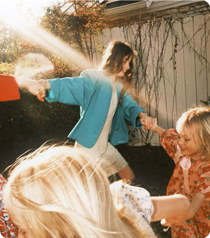 Children and a woman holding hands, playing outdoors in sunlight.