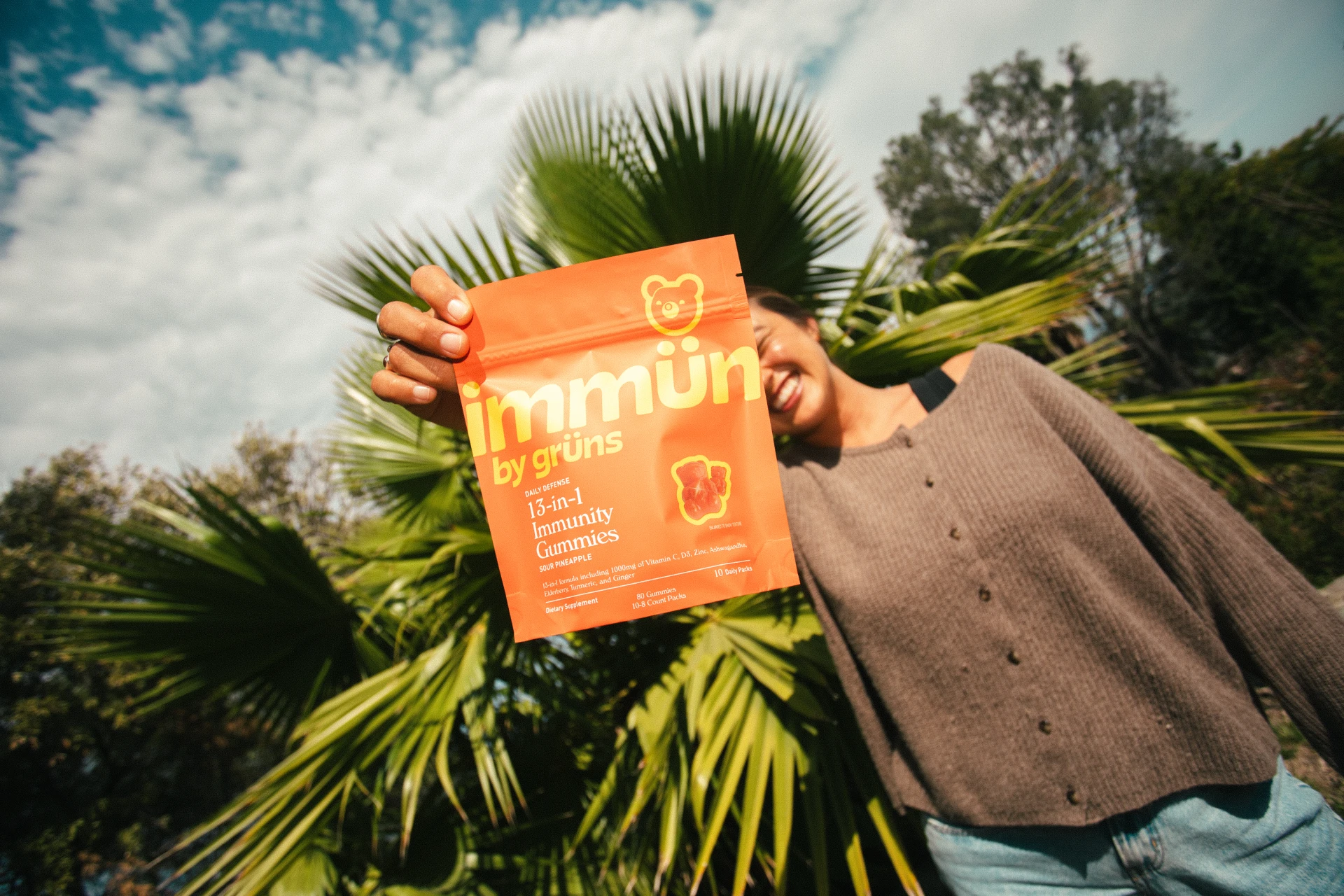 Person holding an orange package of immunity gummies in front of palm leaves.