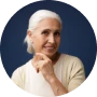 A circular headshot of an older woman with white hair, hand to her chin, against a blue background.