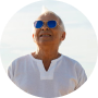 A smiling older person with short white hair, wearing blue sunglasses and a white shirt.