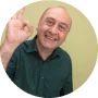 A smiling bald man in a dark green shirt making an okay gesture with his hand.